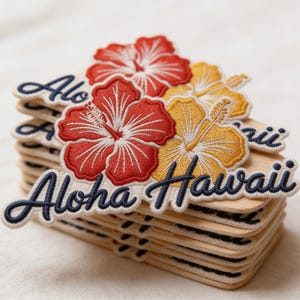 May include: A stack of wooden patches with embroidered hibiscus flowers in red and yellow. The patches have the words "Aloha Hawaii" in navy blue script.