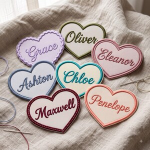 May include: Seven heart-shaped fabric patches with embroidered borders and names. The names Grace, Oliver, Eleanor, Ashton, Chloe, Maxwell, and Penelope are stitched in cursive script on various pastel and solid colored backgrounds with matching thread.