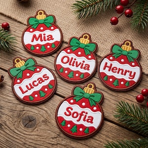 May include: Five circular embroidered patches shaped like Christmas ornaments, each featuring a red and white design with a green bow, a gold cap, and small green pine trees. The names Mia, Olivia, Henry, Lucas, and Sofia are stitched in red.