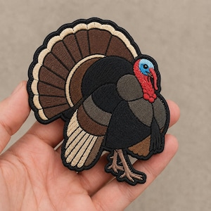 May include: An embroidered patch shaped like a turkey with a fanned tail. The turkey features a black body, brown and tan tail feathers, a bright blue head, and a red wattle. The entire design is outlined in a thick black border with stitching.