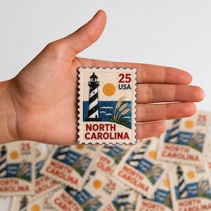 May include: A rectangular embroidered patch shaped like a postage stamp featuring a black and white lighthouse, a yellow sun, blue water, and green grass. The text reads 25 USA and NORTH CAROLINA in red, with a scalloped edge border.