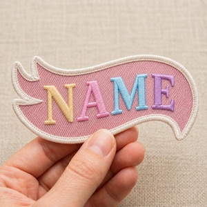 May include: A pink embroidered patch with the word "NAME" in pastel colors. The patch has a white border and a decorative shape. The letters are yellow, pink, blue, and purple.