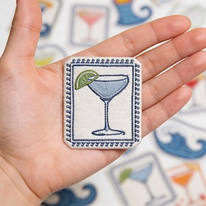 May include: Embroidered patch featuring a blue cocktail with a lime wedge, set within a rectangular frame with a decorative border. The patch is on a white background, with other cocktail-themed patches visible.