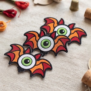 May include: Three embroidered patches featuring a central green eyeball with a black pupil, surrounded by orange and red bat-like wings with black outlines. The patches are arranged in a diagonal line, showcasing detailed stitching and vibrant colors.