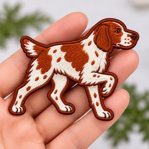 May include: An embroidered iron-on patch shaped like a dog with a white coat and rust-colored spots. The dog is depicted in a standing position with one front paw lifted, featuring detailed stitching that defines its ears, tail, and facial features.