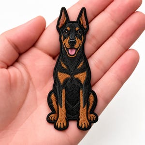 May include: An embroidered iron-on patch featuring a Doberman Pinscher dog sitting in an upright position. The dog has black fur with tan markings on its face, chest, and legs, with pointed ears and a pink tongue visible in its open mouth.