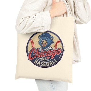 May include: Cream-colored tote bag featuring a vintage-style Chicago Cubs baseball logo. The logo includes a blue bear wearing a cap, with the words "Chicago" and "Baseball" in red and blue. The bag is held by a person.