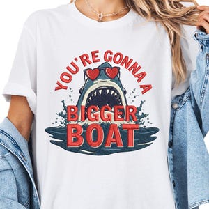 Könnte beinhalten: Weißes T-Shirt mit einem Hai, der eine Herzbrille trägt, mit dem Text "YOU'RE GONNA A BIGGER BOAT" in Rot. Der Hai befindet sich in blauem Wasser mit Spritzern. Ein lustiges, lässiges Design.