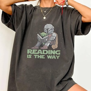 Puede incluir: Camiseta gris oscuro con un gr&aacute;fico de un personaje mandaloriano leyendo un libro con Grogu. El texto "READING IS THE WAY" est&aacute; impreso debajo de la ilustraci&oacute;n. La palabra "PEPPER" est&aacute; impresa en la parte inferior.