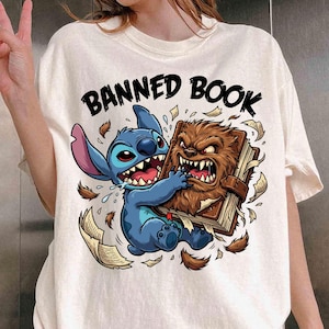 May include: Cream-colored t-shirt featuring a cartoon graphic of a blue alien character and a brown monster fighting over a book. The text "BANNED BOOK" is arched above the characters in black. The shirt is a casual style.