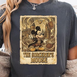 May include: Dark gray t-shirt featuring a vintage-style graphic of Mickey Mouse as a sorcerer. The graphic includes a castle, keys, books, and the text "The Sorcerer's Mouse". The shirt has a crew neck and short sleeves.