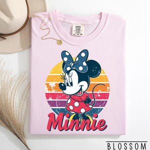 May include: A light pink Comfort Colors t-shirt featuring a Minnie Mouse graphic. The graphic includes Minnie in a red polka dot dress and a blue polka dot bow, set against a retro sunset design. The word "Minnie" is written in red script.