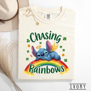 May include: Ivory t-shirt with a cartoon character with angel wings and a halo, lying on a rainbow. The shirt has the text "Chasing Rainbows" in green, with shamrocks and gold coins. A hat and necklace are in the background.