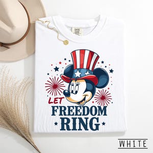 May include: White t-shirt featuring a patriotic Minnie Mouse design. Minnie Mouse is wearing a red, white, and blue Uncle Sam hat. The shirt has the text "LET FREEDOM RING" in navy blue with red star accents. The shirt is labeled "WHITE".