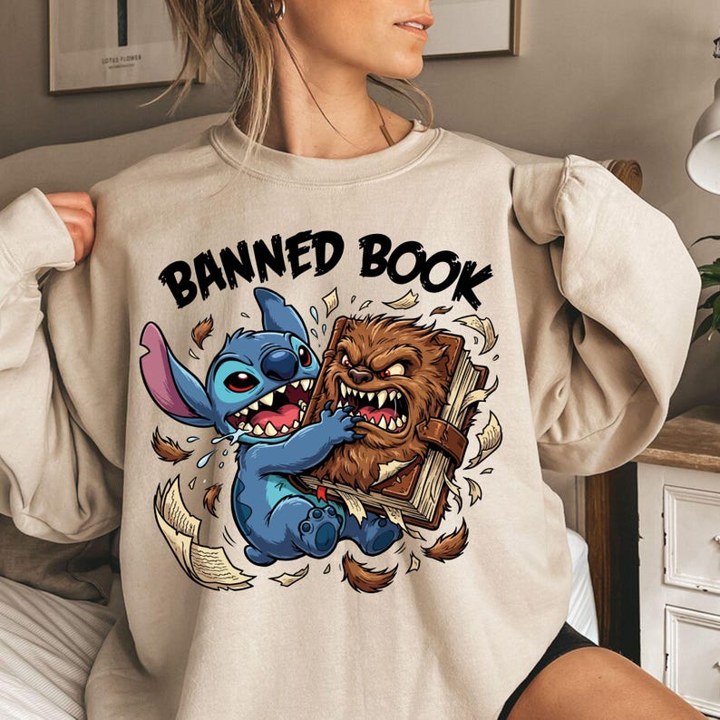 May include: Beige sweatshirt with the text "BANNED BOOK" above a cartoon illustration of a blue creature and a brown creature fighting over a book. The blue creature has large ears and the brown creature has sharp teeth.