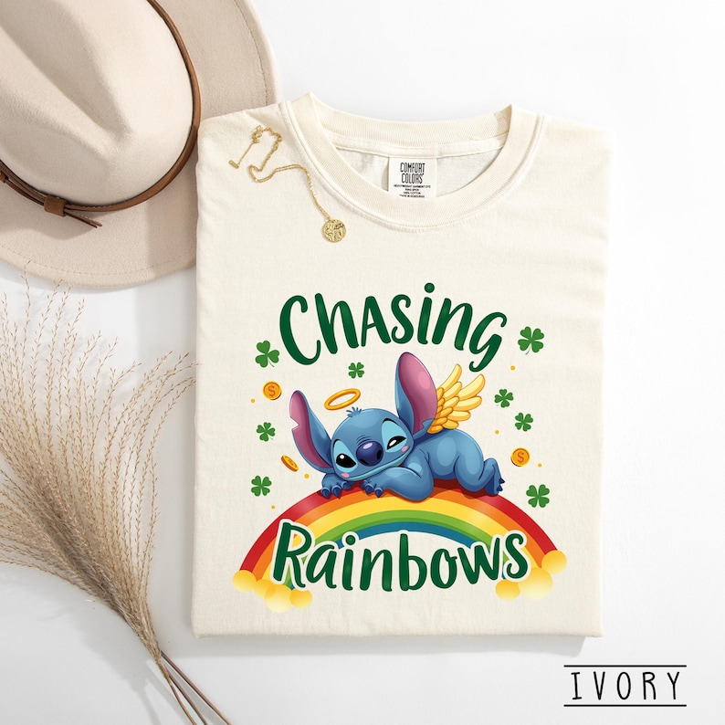 May include: Ivory t-shirt with a cartoon character with angel wings and a halo, lying on a rainbow. The shirt has the text "Chasing Rainbows" in green, with shamrocks and gold coins. A hat and necklace are in the background.