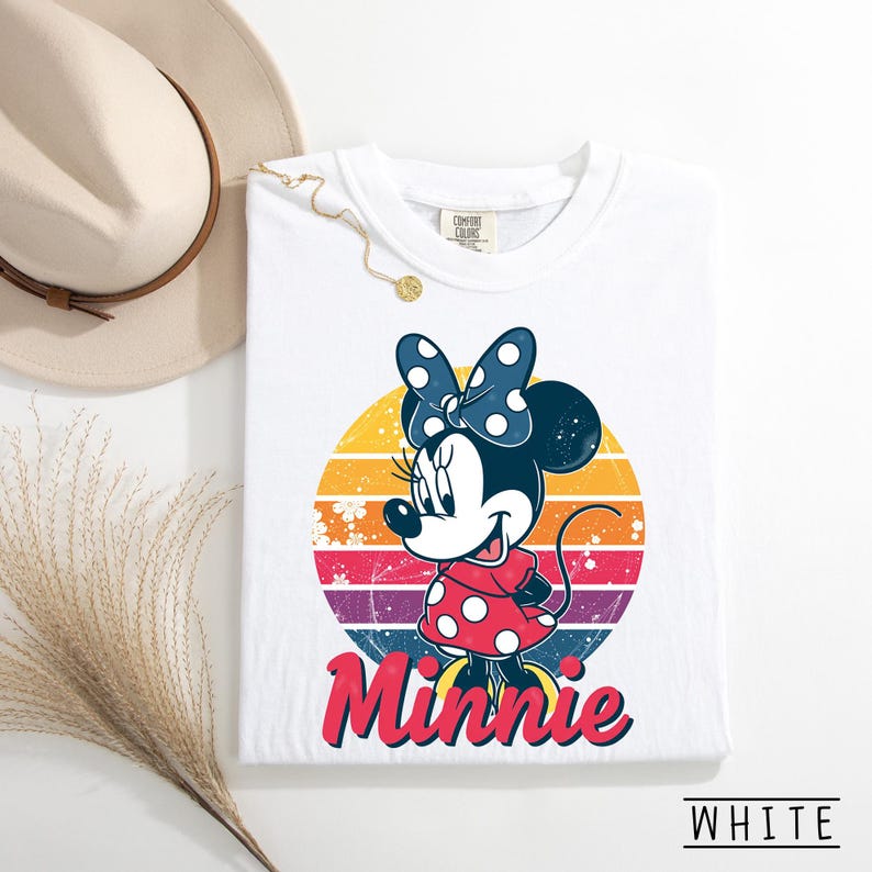 May include: White t-shirt featuring a graphic of Minnie Mouse in a red polka dot dress and blue polka dot bow. The design includes a retro sunset background with the name "Minnie" in red. The shirt is labeled "WHITE" at the bottom.
