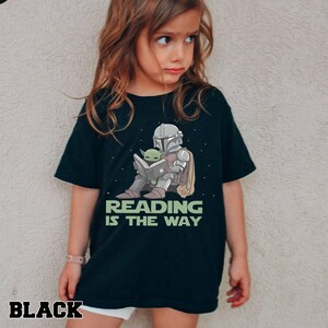 Puede incluir: Camiseta negra con un gr&aacute;fico de un personaje con armadura plateada leyendo un libro con un extraterrestre verde. El texto "READING IS THE WAY" est&aacute; debajo del gr&aacute;fico. La palabra "BLACK" est&aacute; en la parte inferior izquierda.