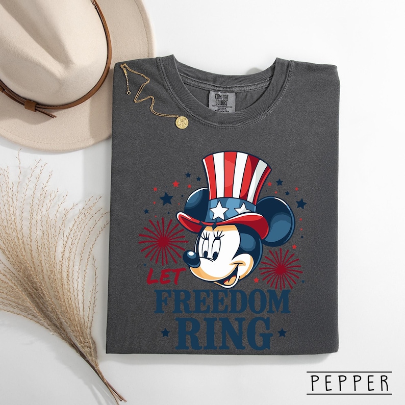May include: Dark gray t-shirt featuring Minnie Mouse in a patriotic Uncle Sam hat. The design includes red, white, and blue stars and fireworks, with the text "LET FREEDOM RING". A gold necklace and a beige hat are visible in the background.