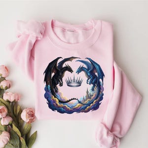 May include: A light pink sweatshirt featuring a fantasy design. Two dragons, one black and one blue, face each other around a silver crown. The dragons are set against a backdrop of colorful clouds and a castle.
