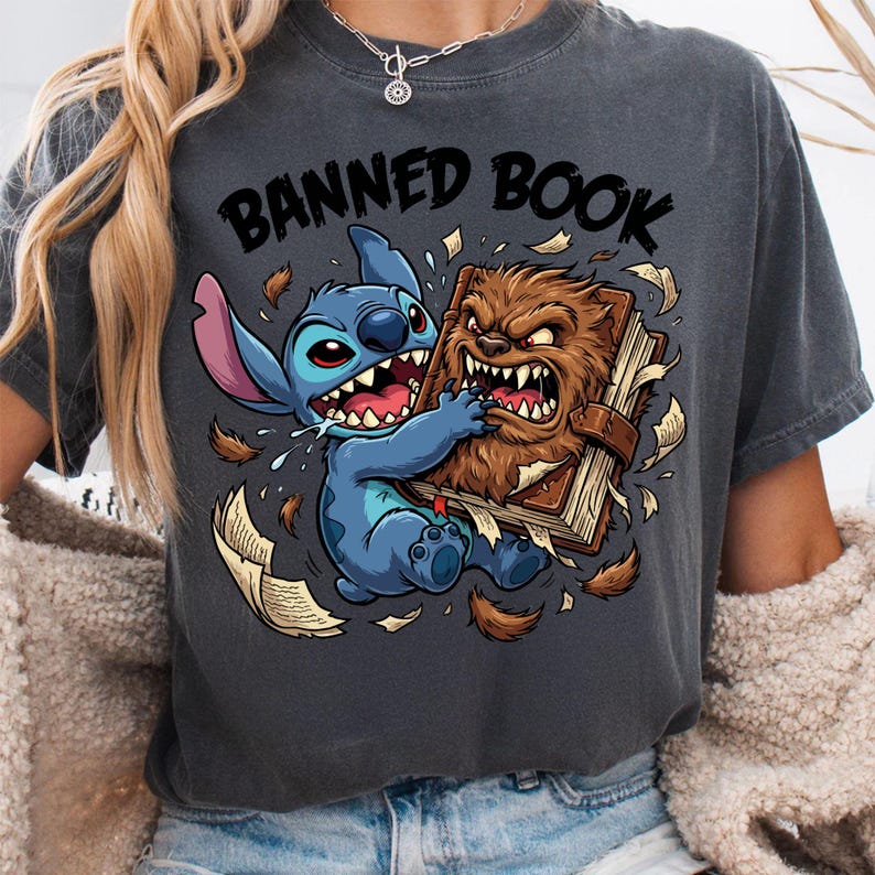 May include: Dark gray t-shirt featuring a cartoon graphic of Stitch from Lilo & Stitch and a furry creature fighting over a book. The text "BANNED BOOK" is printed above the graphic in black.