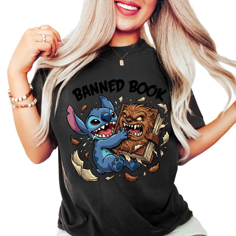 May include: Black t-shirt featuring a cartoon graphic of a blue alien character and a brown furry creature fighting over a book. The text "BANNED BOOK" is arched above the characters.