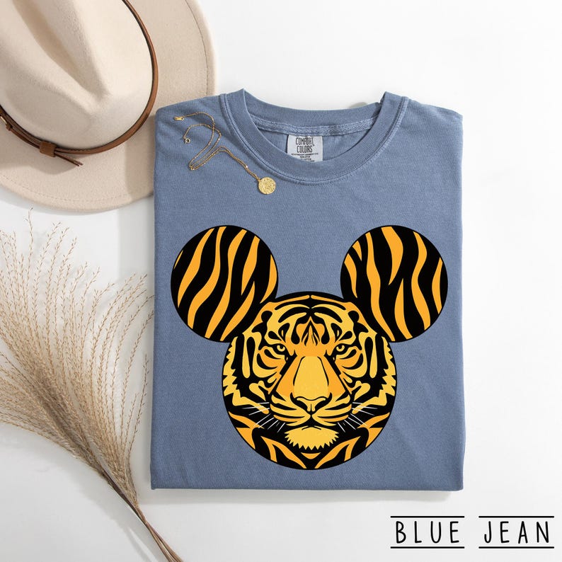 May include: A blue t-shirt with a tiger design. The tiger's face is in the center, with two circular ears on either side. The ears have black and yellow stripes. The text "BLUE JEAN" is at the bottom.