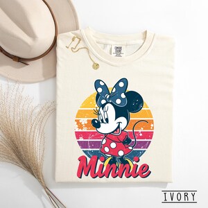 May include: Ivory-colored t-shirt featuring a retro design of Minnie Mouse. Minnie is depicted in a red polka-dot dress and a blue polka-dot bow, set against a colorful sunset background. The word "Minnie" is written in red script below the character.
