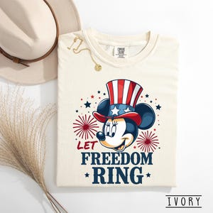 May include: Ivory-colored t-shirt featuring a patriotic Minnie Mouse design. Minnie Mouse is wearing a red, white, and blue Uncle Sam hat. The shirt has the text "LET FREEDOM RING" with star accents. A gold necklace and a beige hat are also visible.