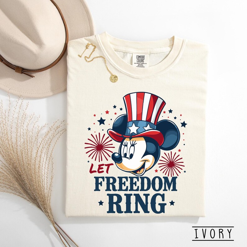 May include: Ivory-colored t-shirt featuring a patriotic Minnie Mouse design. Minnie Mouse is wearing a red, white, and blue Uncle Sam hat. The shirt has the text "LET FREEDOM RING" with star accents. A gold necklace and a beige hat are also visible.