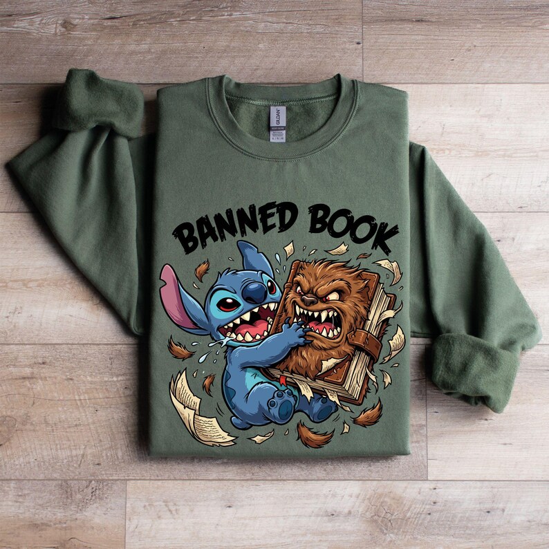 May include: Olive green sweatshirt featuring a graphic of a blue cartoon character and a brown furry creature fighting over a book. The text "BANNED BOOK" is above the characters. The sweatshirt has a crew neck and long sleeves.
