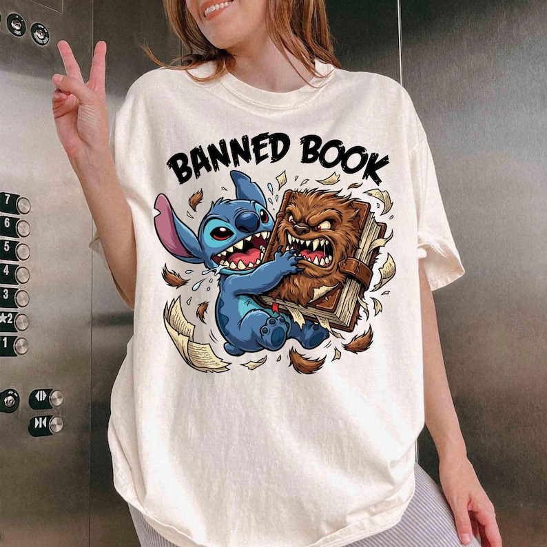 May include: Cream-colored t-shirt featuring a cartoon graphic of a blue alien character and a brown monster fighting over a book. The text "BANNED BOOK" is arched above the characters in black. The shirt is a casual style.