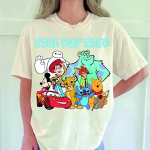 Może przedstawiać: Kremowy t-shirt z kr&oacute;tkim rękawem z napisem COOL BOY CLUB jasnoniebieskimi literami nad grafiką przedstawiającą r&oacute;żne postacie animowane, w tym Baymaxa, Chudego, Myszkę Miki, Zygzaka McQueena, Simbę, Nicka Bajera, Sulleya, Sticha, Kubusia Puchatka i Remy'ego.