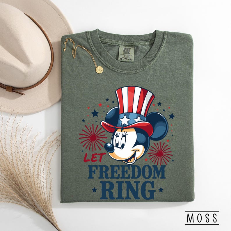 May include: Olive green Comfort Colors t-shirt featuring Minnie Mouse in a patriotic Uncle Sam hat. The shirt has the text "LET FREEDOM RING" in blue, with red star and firework accents. A gold necklace and a beige hat are also in the image.