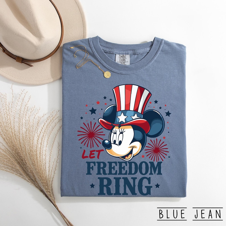 May include: Blue jean colored t-shirt featuring Minnie Mouse in a patriotic top hat. The shirt has the text "LET FREEDOM RING" in red and blue. A hat and jewelry are visible in the background.