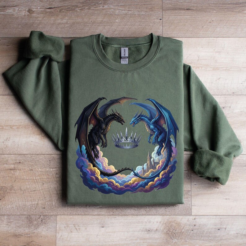 May include: Olive green sweatshirt featuring a fantasy design. Two dragons, one black and one blue, face each other around a silver crown. The dragons are set against a backdrop of colorful clouds and a cityscape.