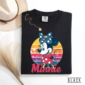 May include: Black sweatshirt featuring a Minnie Mouse graphic. The graphic shows Minnie in a red polka dot dress and a blue polka dot bow, set against a retro sunset design. The word "Minnie" is written in red script. The sweatshirt is labeled "BLACK".