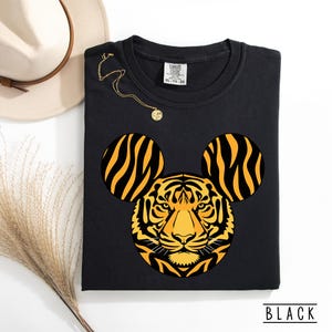 May include: Black sweatshirt featuring a tiger design with Mickey Mouse ears. The tiger's face is yellow and orange with black stripes. The word "BLACK" is printed at the bottom. A gold necklace and a beige hat are also in the image.