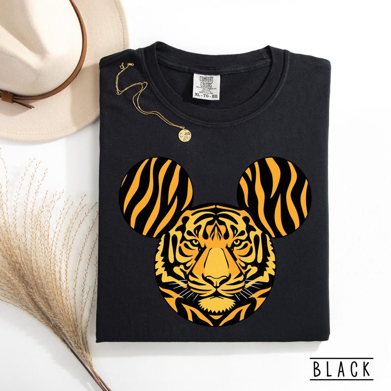 May include: Black sweatshirt featuring a tiger design with Mickey Mouse ears. The tiger's face is yellow and orange with black stripes. The word "BLACK" is printed at the bottom. A gold necklace and a beige hat are also in the image.
