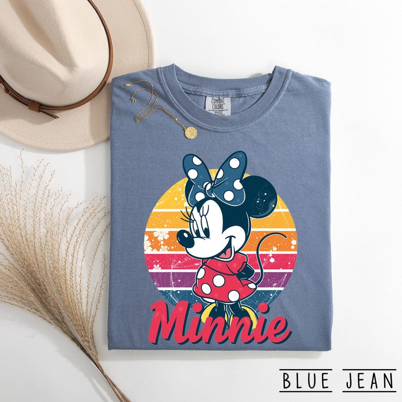 May include: Blue jean colored t-shirt featuring a Minnie Mouse graphic. The graphic includes Minnie in a red polka dot dress and a blue polka dot bow, set against a colorful sunset design. The word "Minnie" is written in red script.