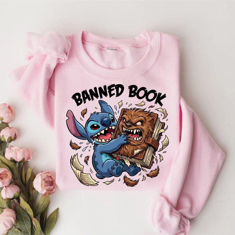 May include: A light pink sweatshirt featuring a cartoon graphic of Stitch from Lilo & Stitch and a brown, furry creature fighting over a book. The text "BANNED BOOK" is printed above the characters. The sweatshirt has long sleeves.