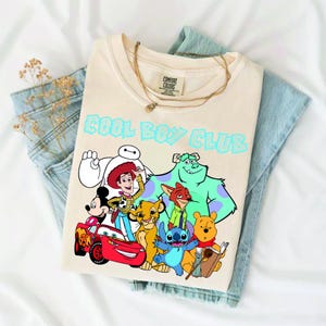 Może przedstawiać: Kremowy t-shirt z napisem COOL BOY CLUB jasnoniebieskimi literami nad grafiką przedstawiającą r&oacute;żne postacie animowane, w tym Myszkę Miki, Chudego, Zygzaka McQueena, Baymaxa, Simbę, Nicka Wilde'a, Sulleya, Sticha i Kubusia Puchatka.