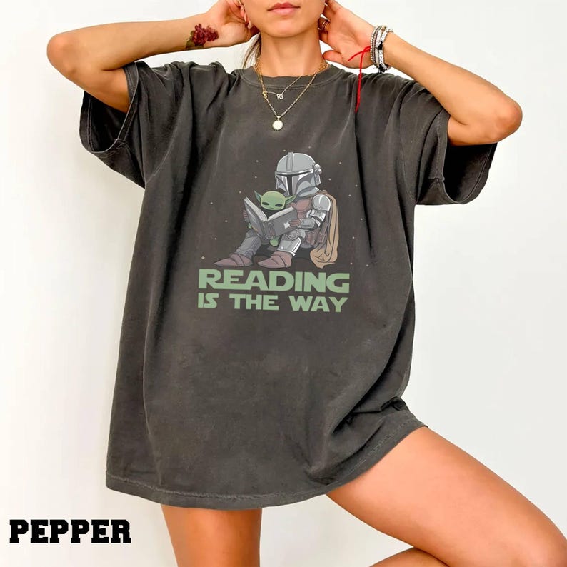 Puede incluir: Camiseta gris oscuro con un gr&aacute;fico de un personaje mandaloriano leyendo un libro con Grogu. El texto "READING IS THE WAY" est&aacute; impreso debajo de la ilustraci&oacute;n. La palabra "PEPPER" est&aacute; impresa en la parte inferior.