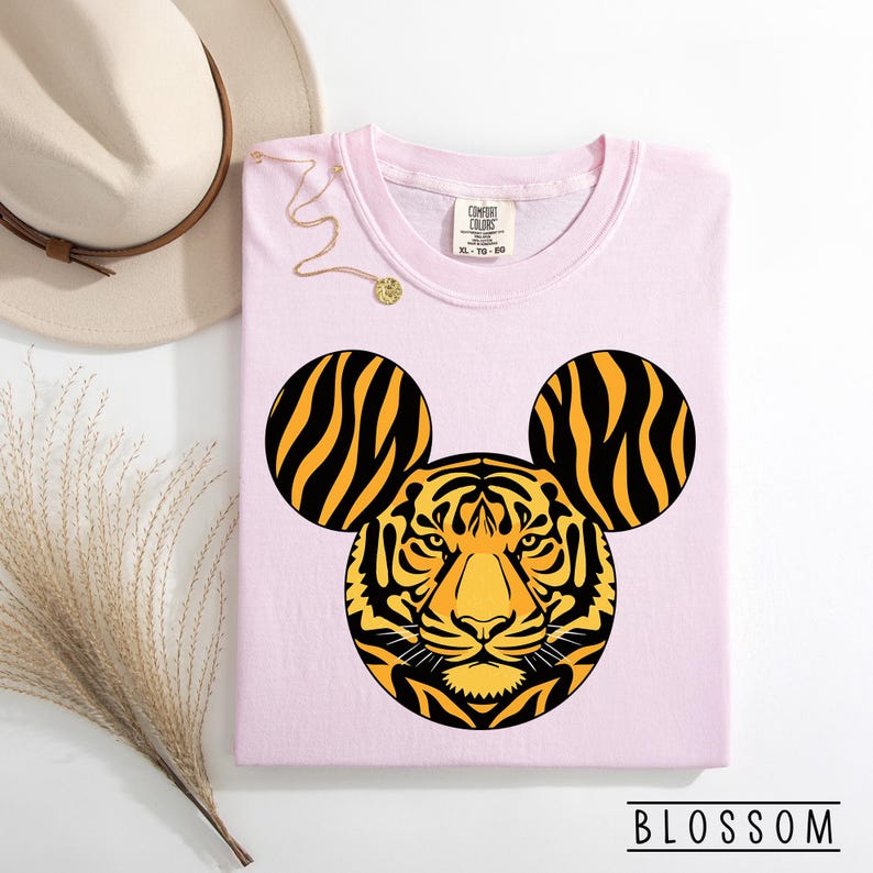 May include: A light pink t-shirt featuring a tiger face design with Mickey Mouse ears. The tiger's face is yellow and black, with black stripes on the ears. The text "BLOSSOM" is at the bottom.