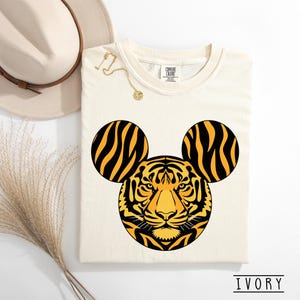 May include: Ivory-colored t-shirt featuring a tiger design with Mickey Mouse ears. The tiger's face is in the center, with black and yellow stripes. The shirt has a crew neck and short sleeves. The word "IVORY" is printed at the bottom.