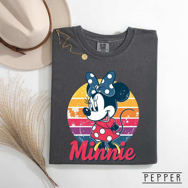 May include: Dark gray t-shirt featuring a Minnie Mouse graphic. Minnie is wearing a red dress with white polka dots and a blue bow with white polka dots. The word "Minnie" is written in red script below the graphic. The shirt is part of the Comfort Colors brand.