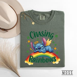 May include: Olive green t-shirt with the text "Chasing Rainbows" and a cartoon character lying on a rainbow. The character has blue fur, pink ears, and golden wings. The shirt also features shamrocks and gold coins.