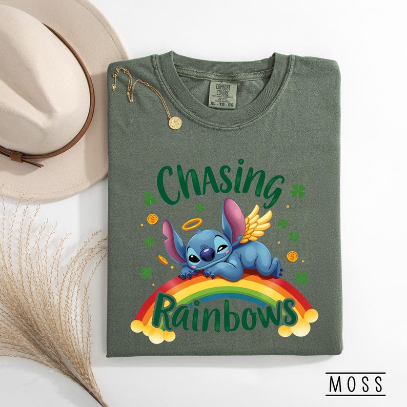 May include: Olive green t-shirt with the text "Chasing Rainbows" and a cartoon character lying on a rainbow. The character has blue fur, pink ears, and golden wings. The shirt also features shamrocks and gold coins.