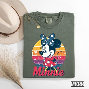 May include: Olive green t-shirt featuring a graphic of Minnie Mouse in a red polka dot dress and blue bow, set against a retro sunset design. The word "Minnie" is written in red script. The shirt is folded, with a hat and necklace visible.