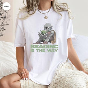 Puede incluir: Camiseta blanca con un gr&aacute;fico de un personaje con armadura leyendo un libro con una peque&ntilde;a criatura verde. El texto "READING IS THE WAY" est&aacute; impreso en verde debajo de la ilustraci&oacute;n. La camiseta tiene un corte holgado y extragrande.